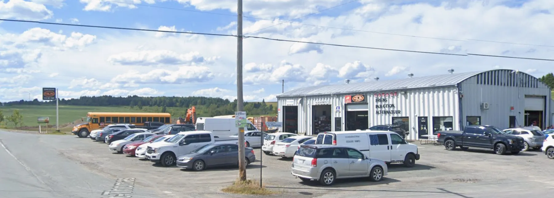 Le Garage Auto HLR vu de la route : le long bâtiment de tôle, ses portes de baie dont une marquée « Alignement », les mots freins, injection et alternateur peints sur la façade, et la cour remplie de véhicules de clients.